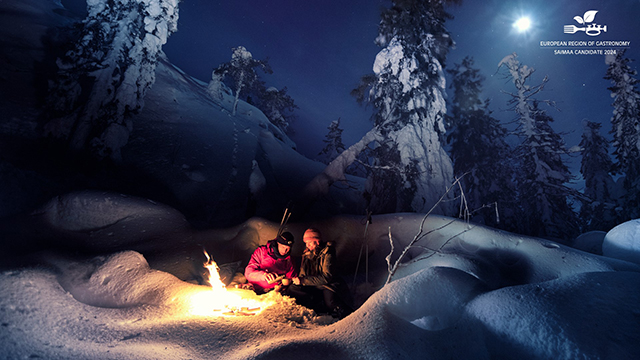 Kaksi ihmistä tulen ääressä metsässä tummana talvi-iltana.